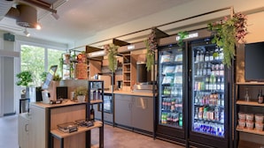 A modern bar area with a coffee machine, a beverage dispenser, and a refrigerator stocked with drinks. - ibis Köln Airport (Cologne)