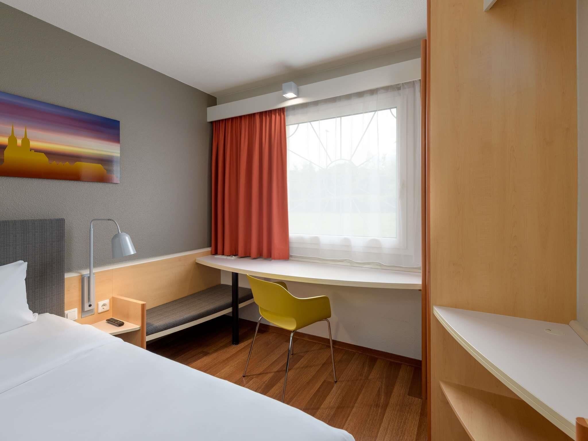 A hotel room with a bed, a desk, a chair, a lamp, and a window with curtains.