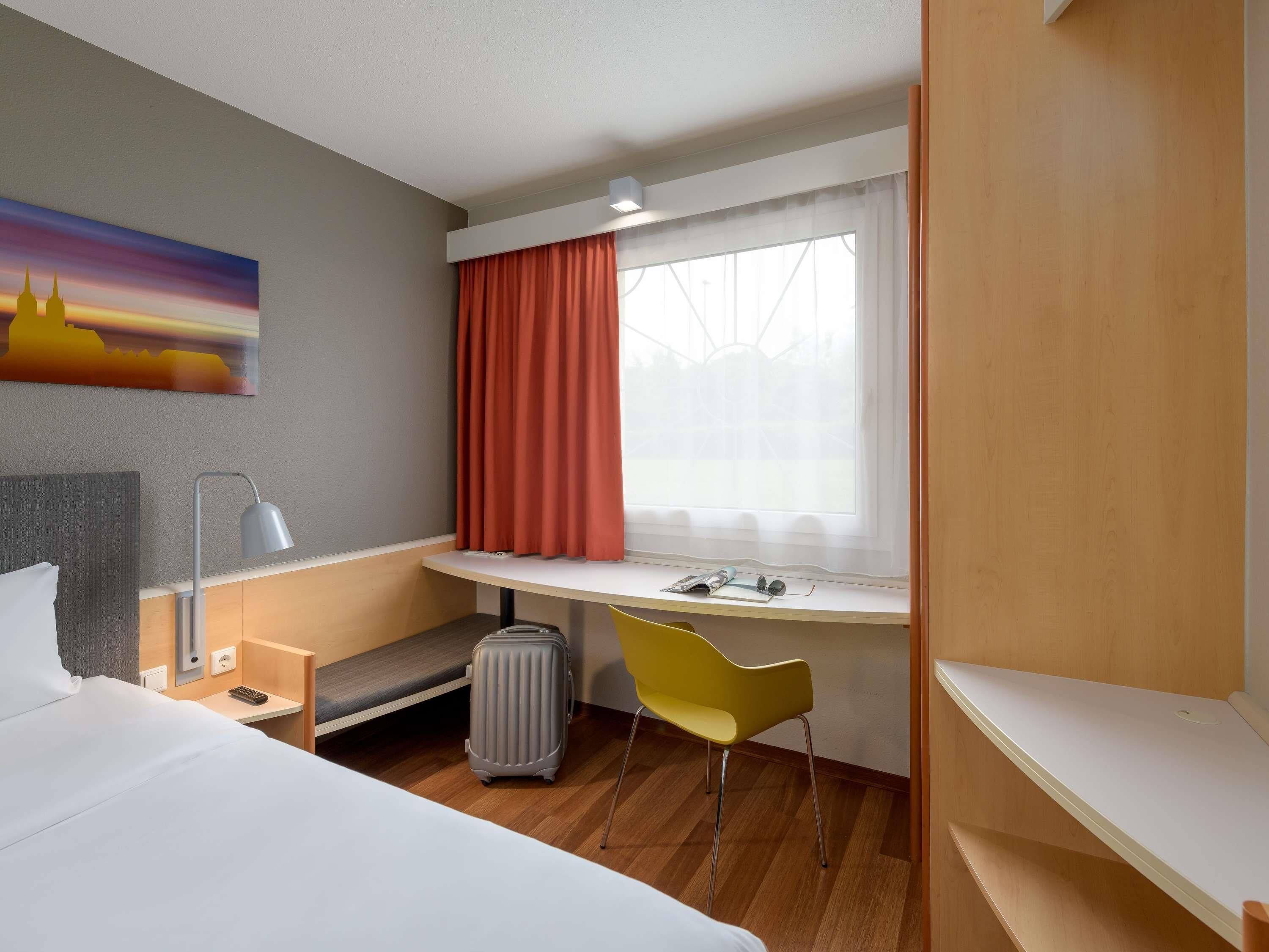 Ein Hotelzimmer mit Bett, Schreibtisch mit Stuhl, Koffer, Lampe und einem Bild an der Wand.