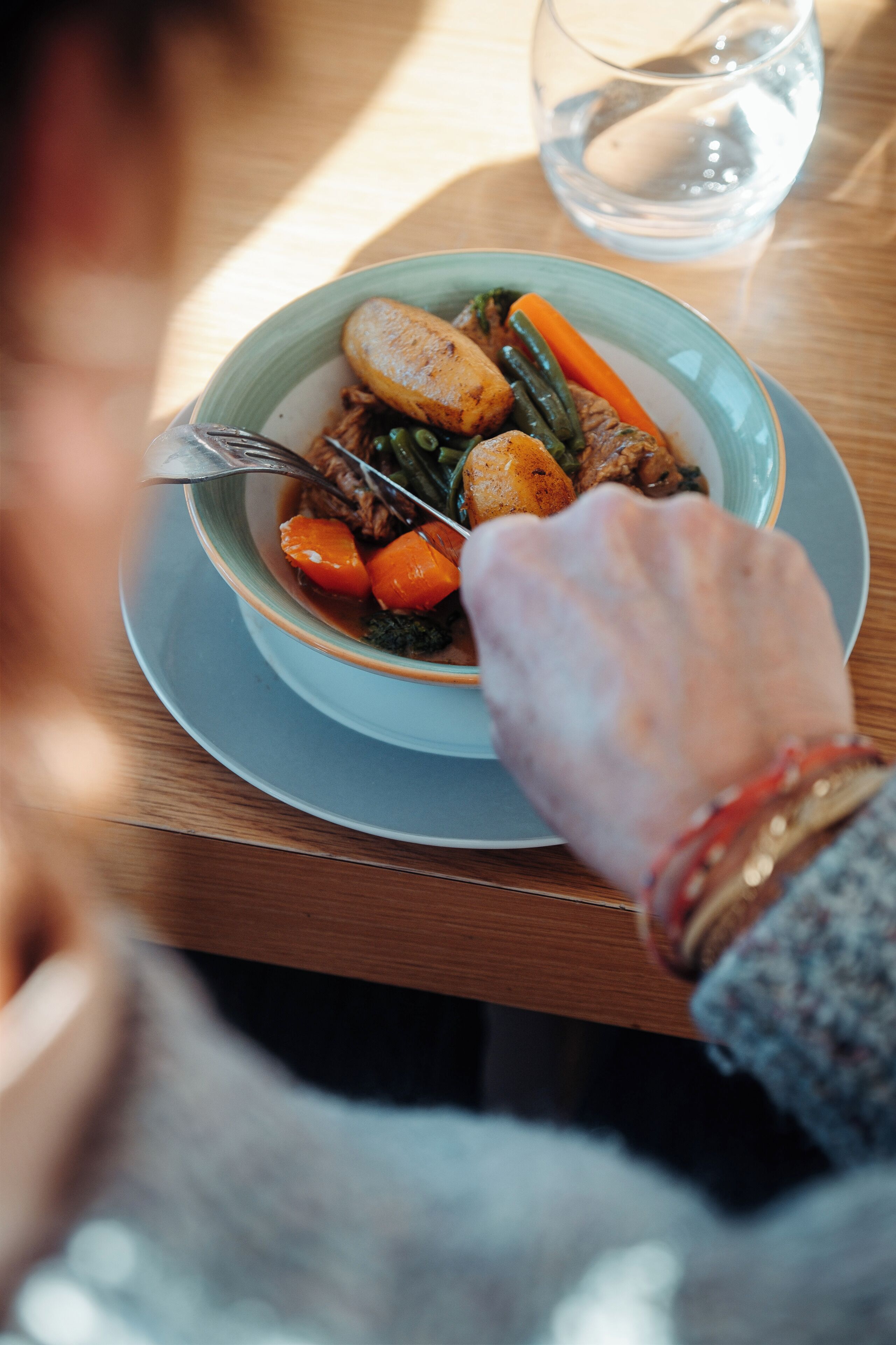 Una mesa de comedor con un plato de comida, un vaso de agua y la mano de una persona alcanzando el plato.