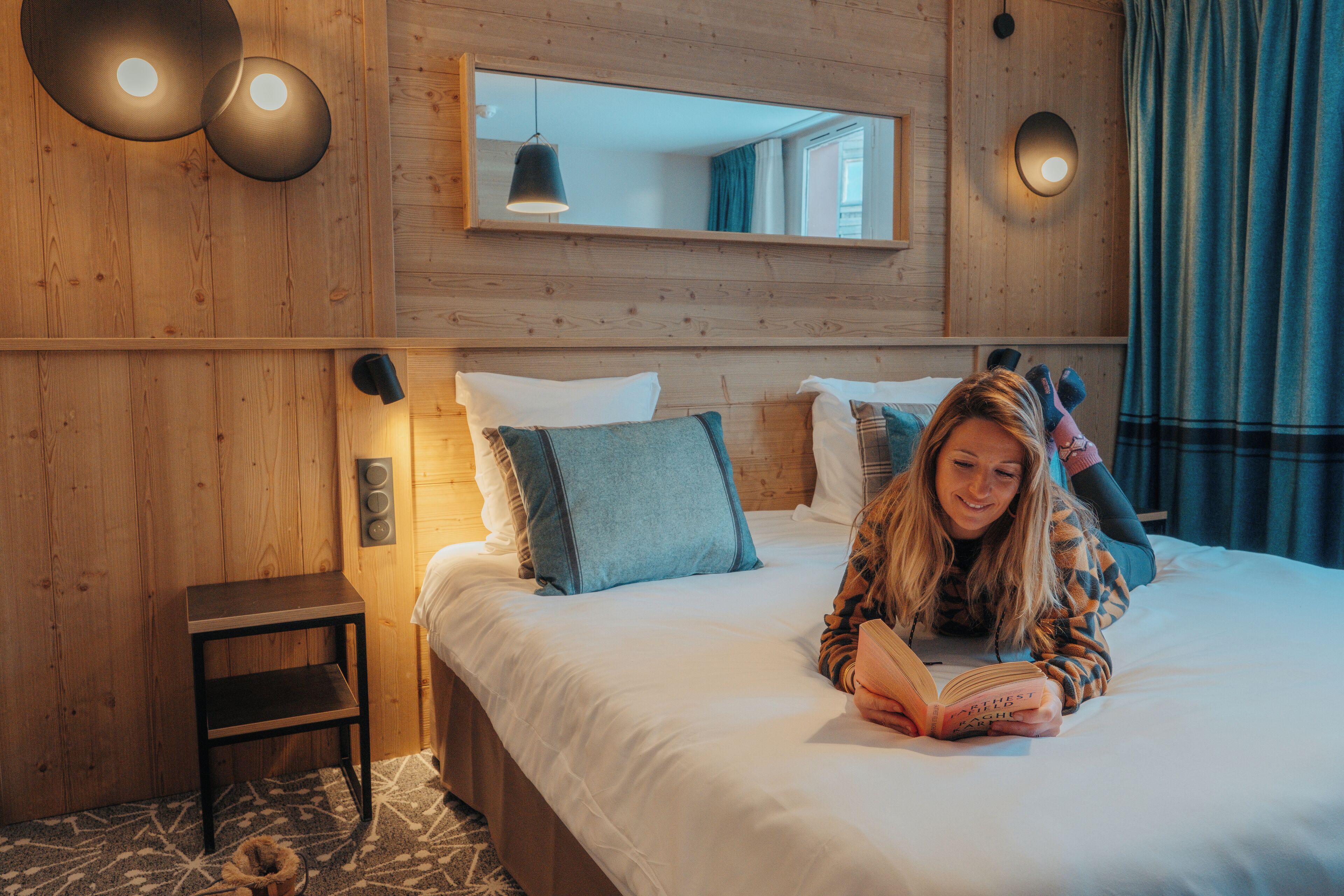 A hotel room with a wooden wall, a bed with white bedding, a bedside table, and a mirror above the bed.