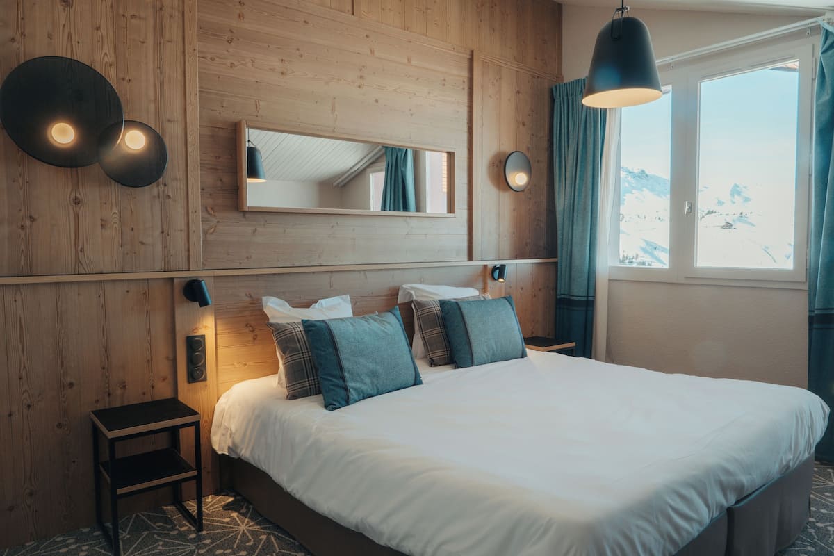 Ein schlafreicher Raum mit Holzwänden, einem großen Bett, zwei Nachttischlampen, einem Spiegel und einem Fenster mit Vorhängen.