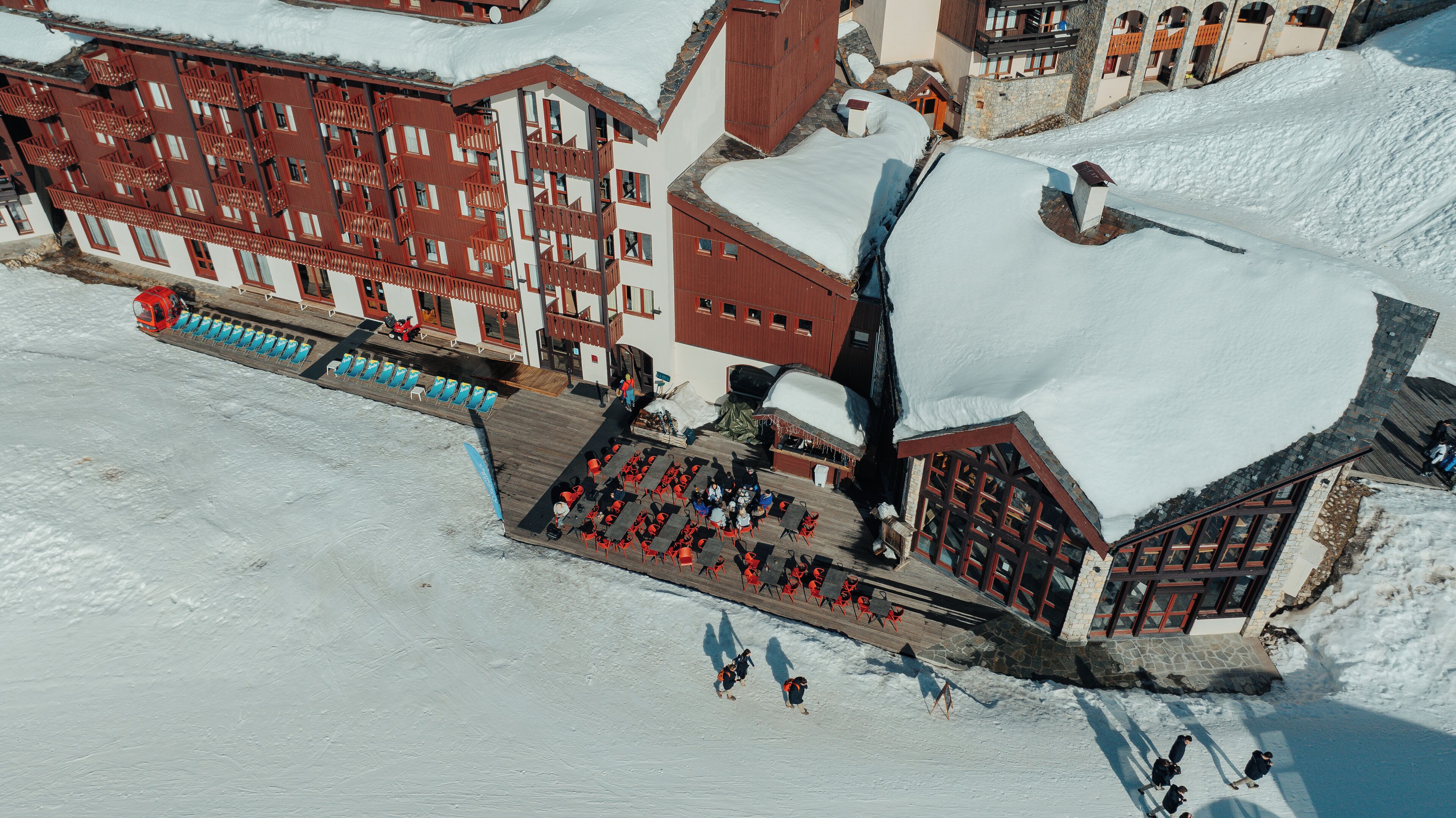 A snow-covered outdoor area with a wooden deck and red chairs, surrounded by buildings with balconies and stone walls.