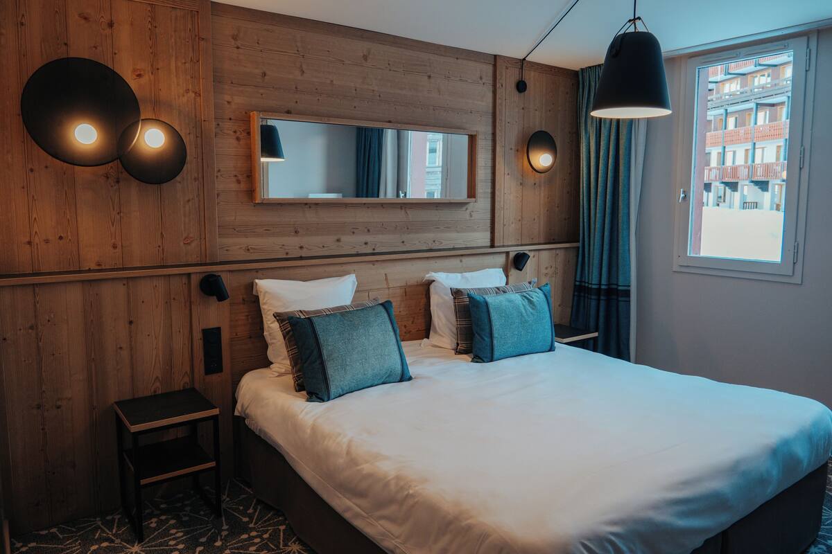 Ein modernes Hotelzimmer mit einer Holzwand, einem großen Bett mit weißer Bettwäsche und türkisfarbenen Kissen, einem kleinen Nachttisch und einem Fenster mit Blick auf Gebäude.