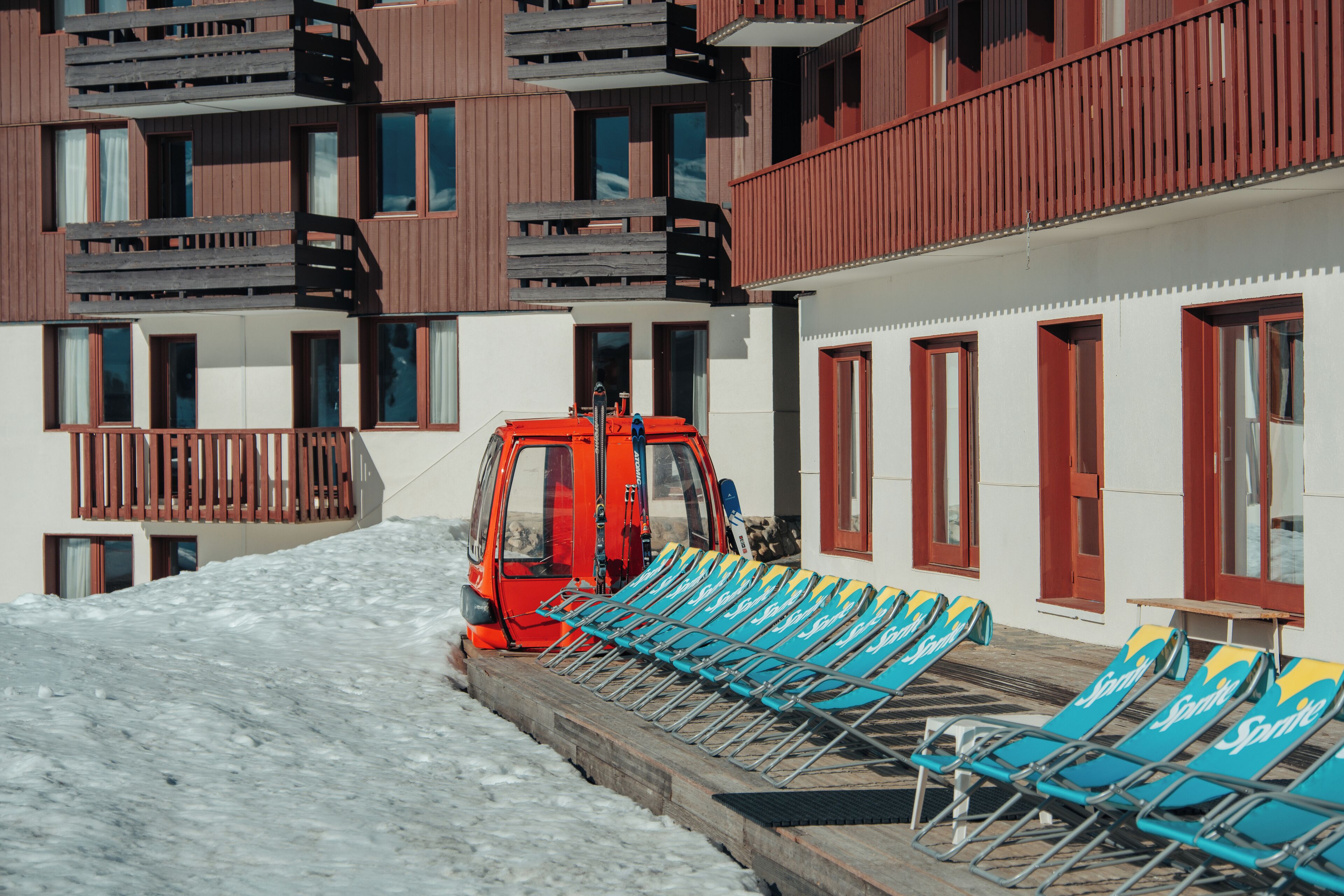 A snow-covered outdoor area with a row of blue lounge chairs and a red vehicle.