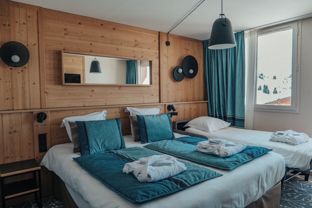 Ein Hotelzimmer mit Holzwandverkleidung, einem großen Fenster mit Blick auf eine verschneite Landschaft, zwei Betten mit türkisblauer Bettwäsche und einem Spiegel über der Kopfteil.