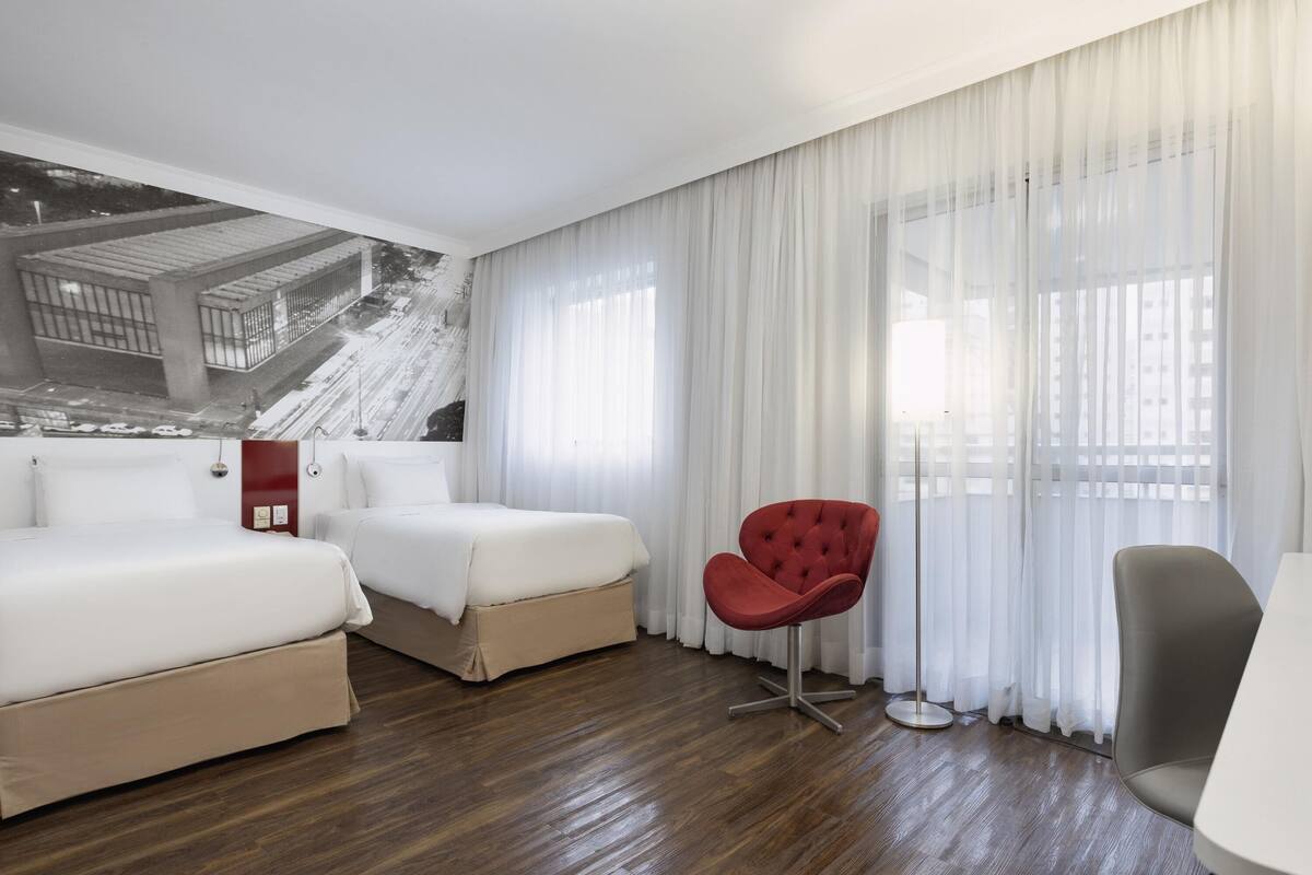 Habitación de hotel con dos camas, una silla roja, una silla gris y un ventanal con cortinas translúcidas.