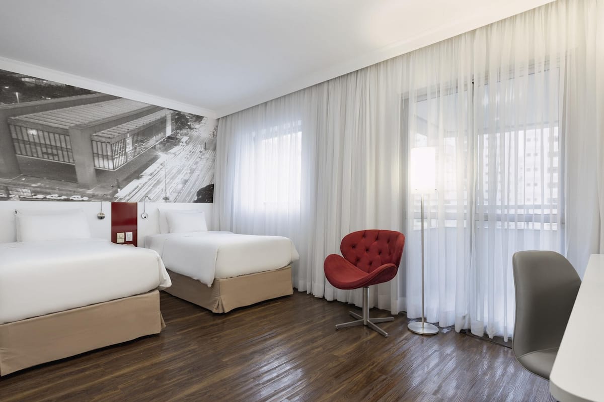 Ein Hotelzimmer mit zwei Betten, einem roten Sessel, einem grauen Sessel und einem groĂen Fenster mit durchsichtigen VorhĂ€ngen.