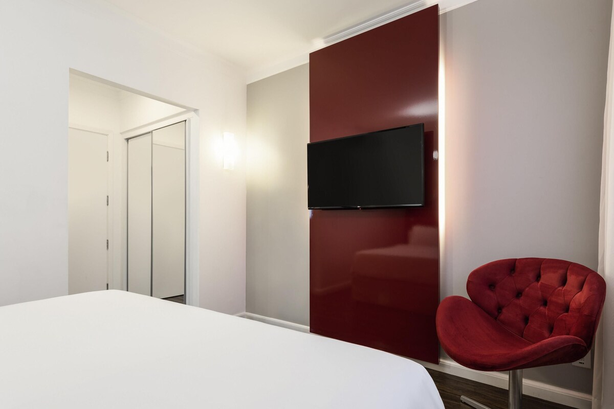 A hotel room with a bed, a red upholstered chair, a television mounted on a red wall, and a door leading to another room.