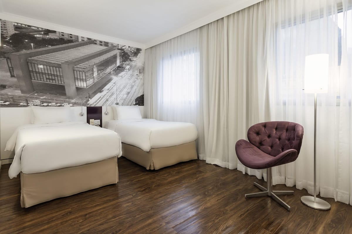 Una habitación de hotel con dos camas, una silla morada y un ventanal con cortinas translúcidas.