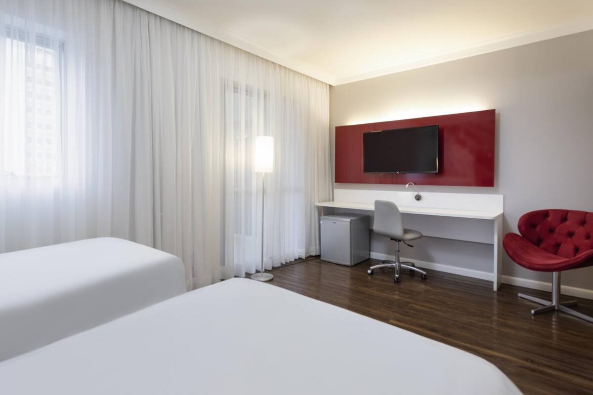 Habitación de hotel con dos camas, un escritorio, una silla, un televisor y una ventana con cortinas.