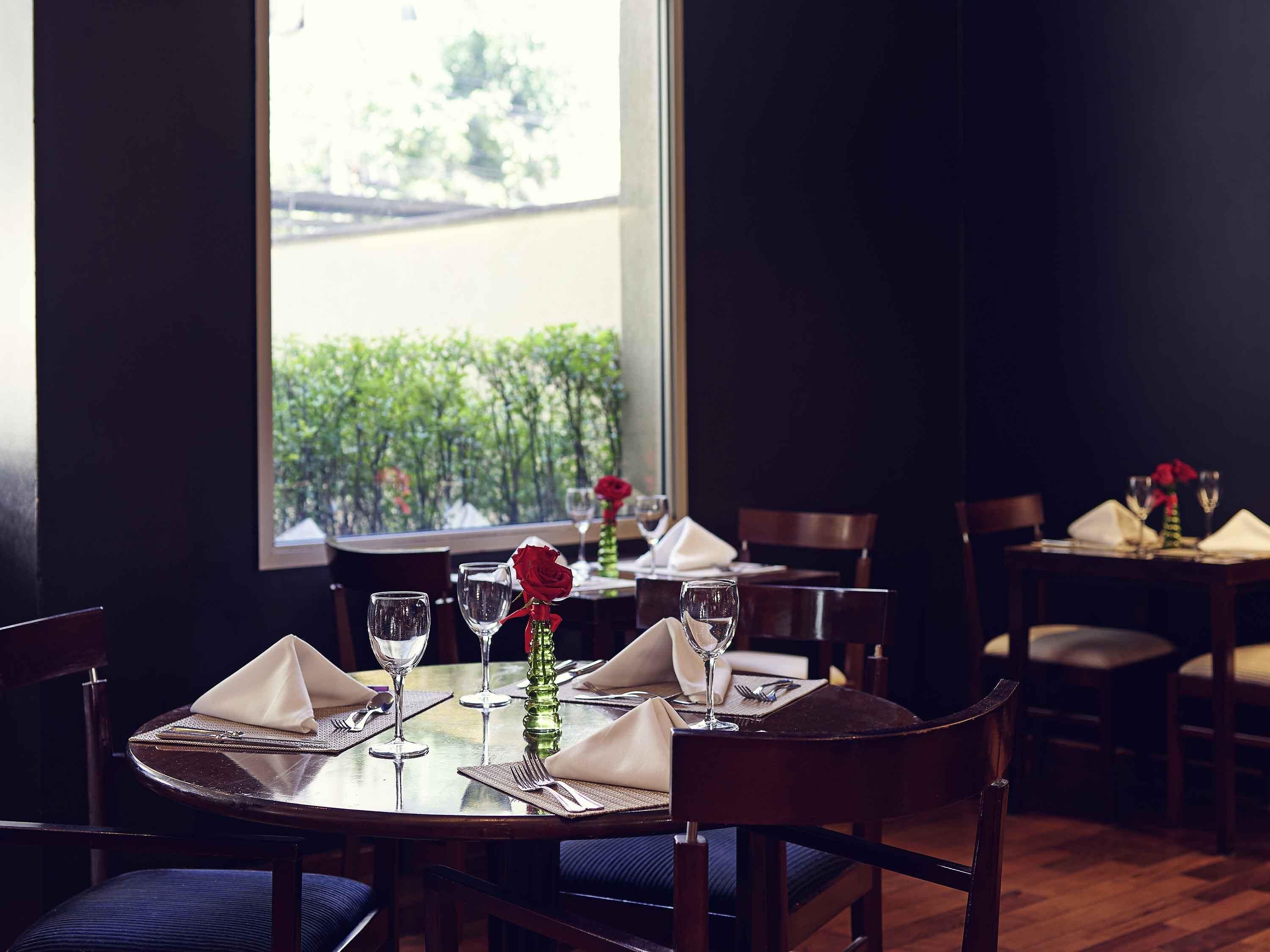 Un comedor con mesas dispuestas para dos, cada una con un jarrón de rosas rojas, copas de vino y cubiertos.