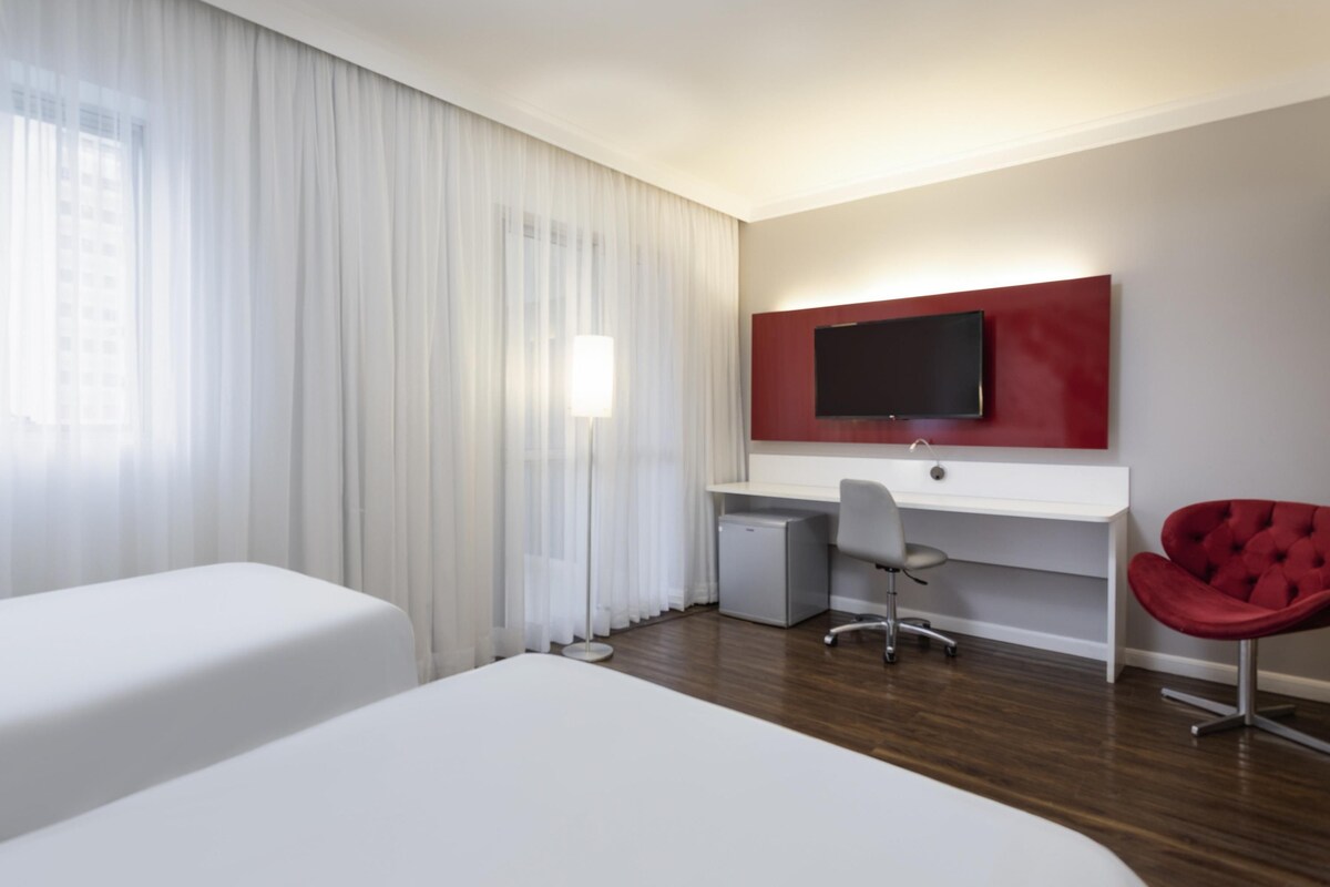 A hotel room with two beds, a desk, a chair, a TV, and a window with curtains.