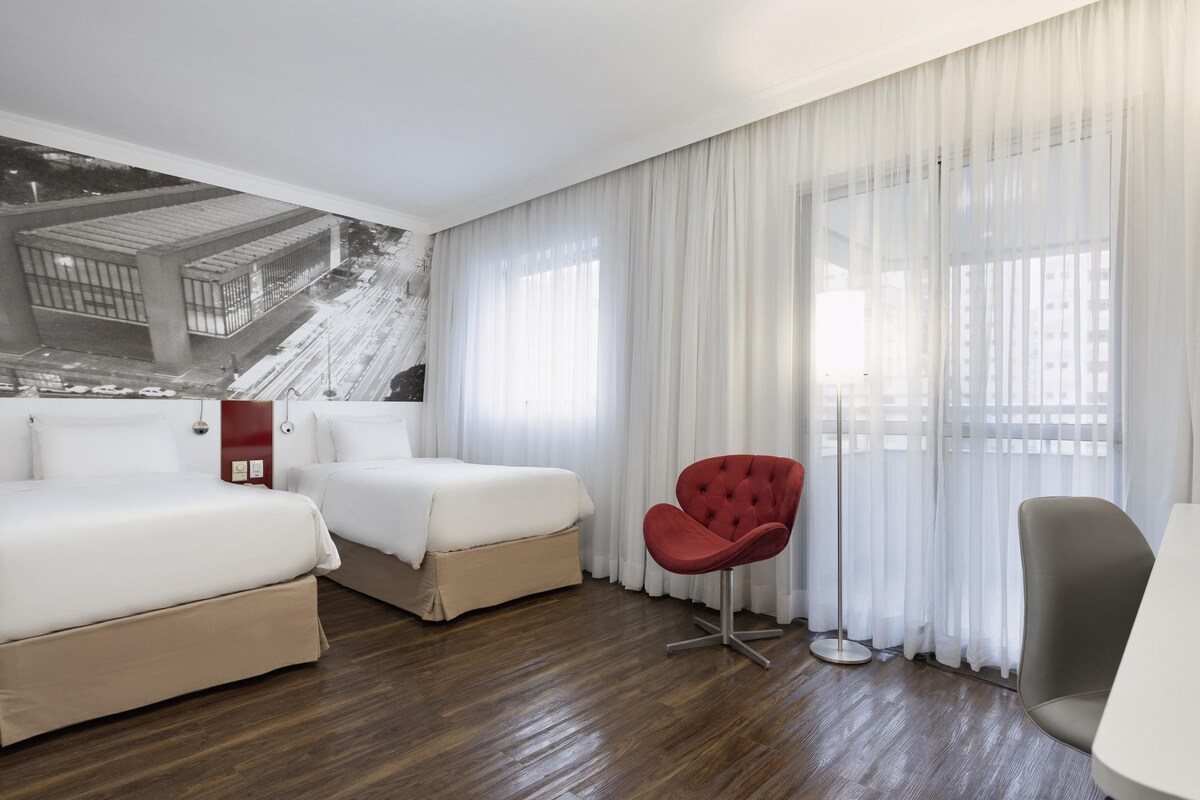 A hotel room with two beds, a red chair, a grey chair, and a large window with sheer curtains.
