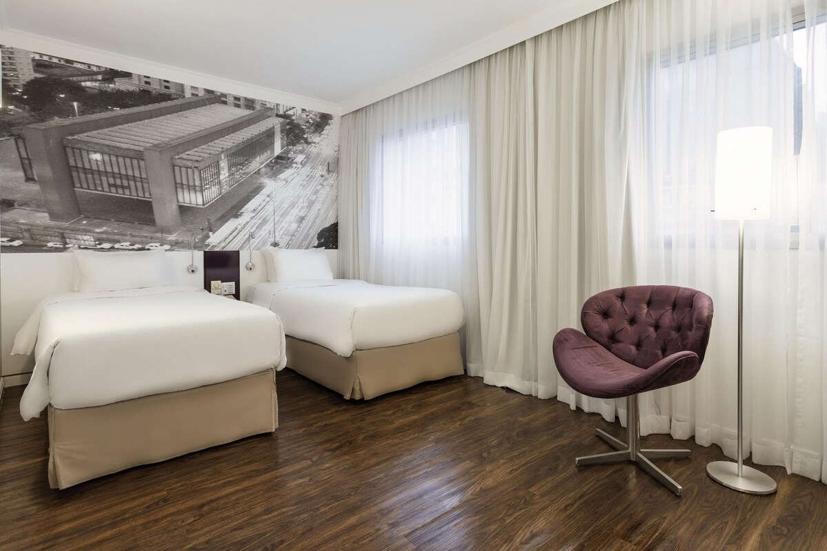 A hotel room with two beds, a purple chair, and a large window with sheer curtains.
