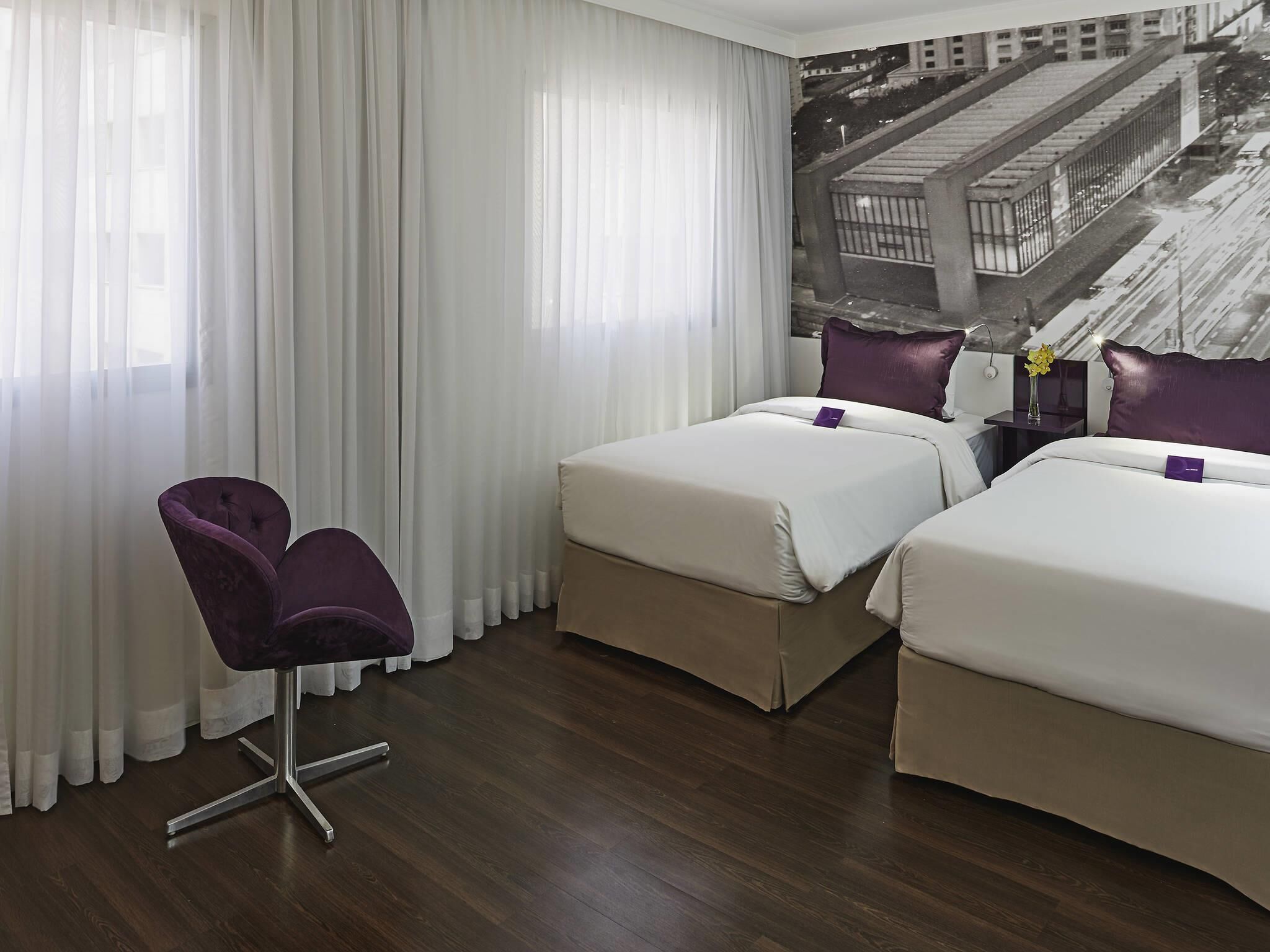 Hotellihuone, jossa on kaksi sänkyä, tuoli, yöpöytä ja suuri ikkuna, jossa on ohuet verhot.