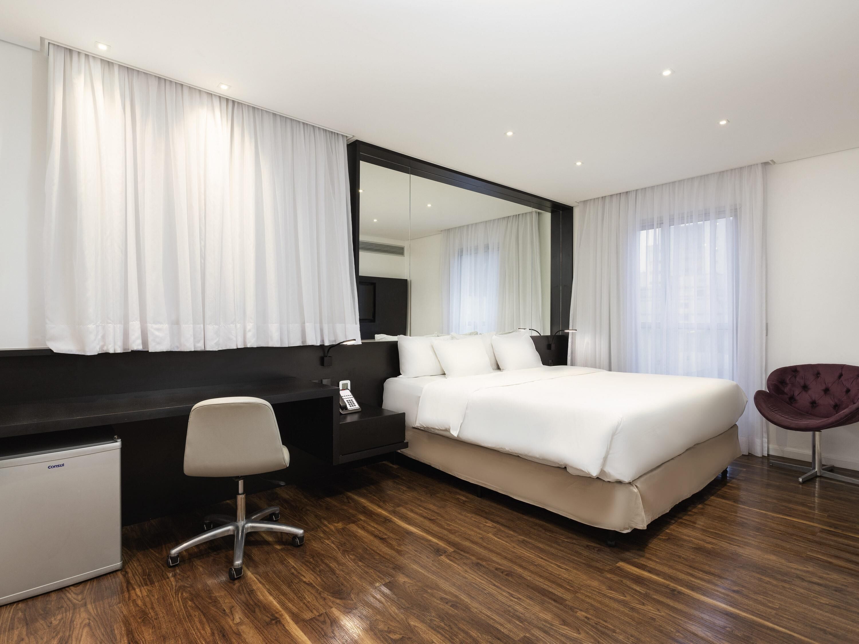 A modern hotel room with a large bed, a desk with a chair, a small fridge, and a large mirror.