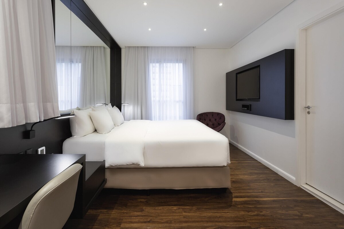 A modern hotel room with a large bed, a desk, a chair, a TV mounted on the wall, and a door.