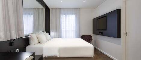A modern hotel room with a large bed, a desk, a chair, a TV mounted on the wall, and a door.