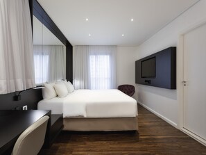 Una habitación de hotel moderna con una cama grande, un escritorio, una silla, un televisor montado en la pared y una puerta.