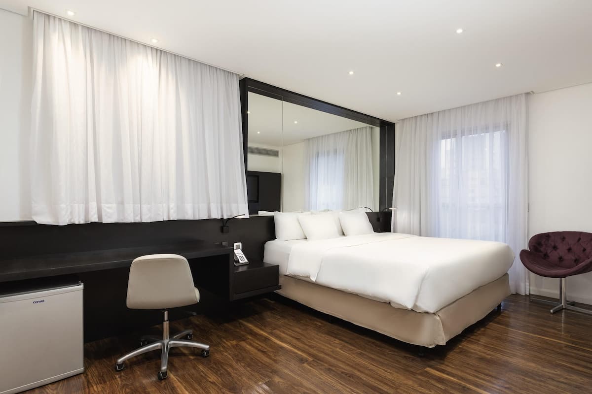 Habitación de hotel moderna con una cama grande, un escritorio con una silla, un refrigerador pequeño y un espejo amplio.