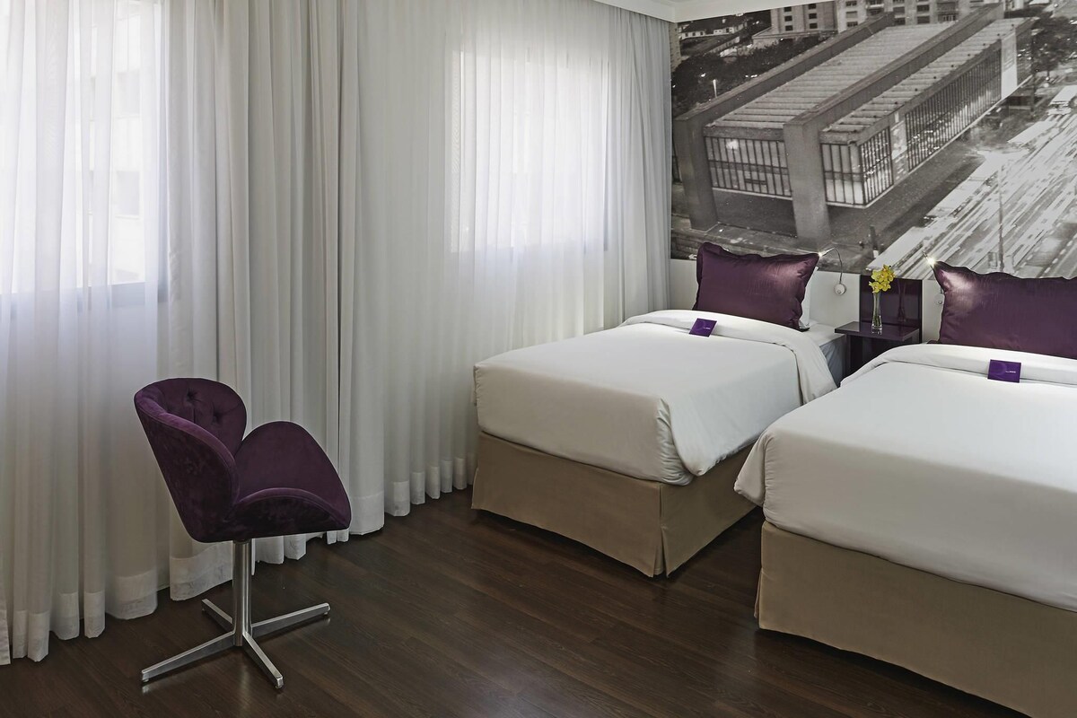 A hotel room with two beds, a chair, a nightstand, and a large window with sheer curtains.
