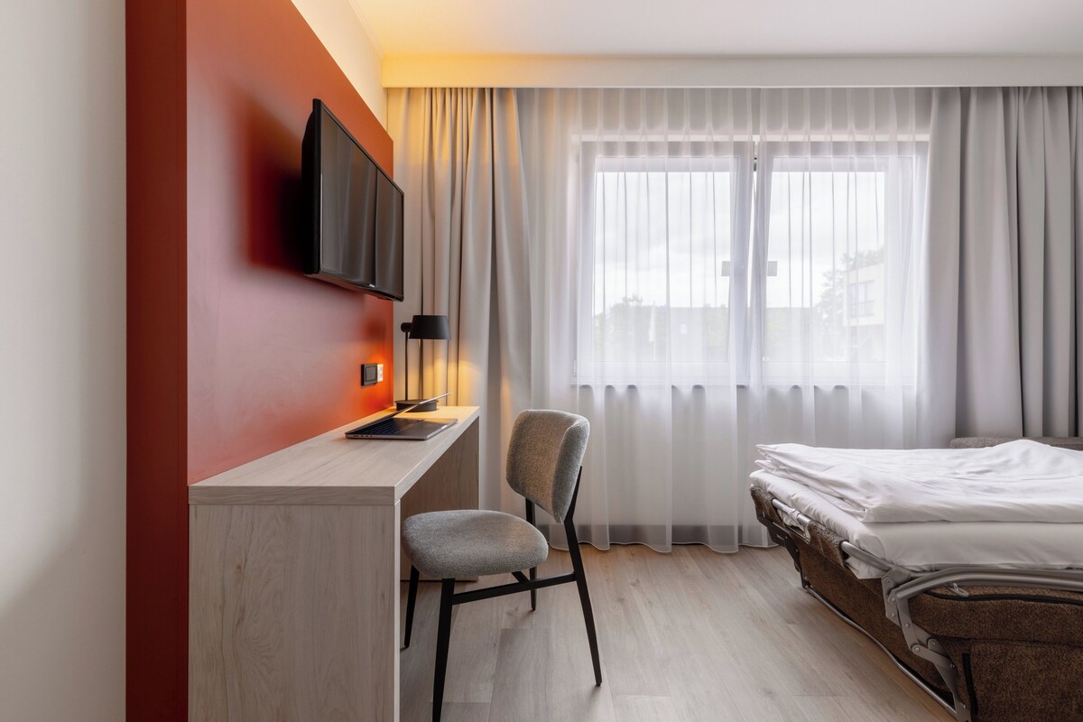 A hotel room with a bed, a desk with a chair, a TV mounted on the wall, and a window with curtains.