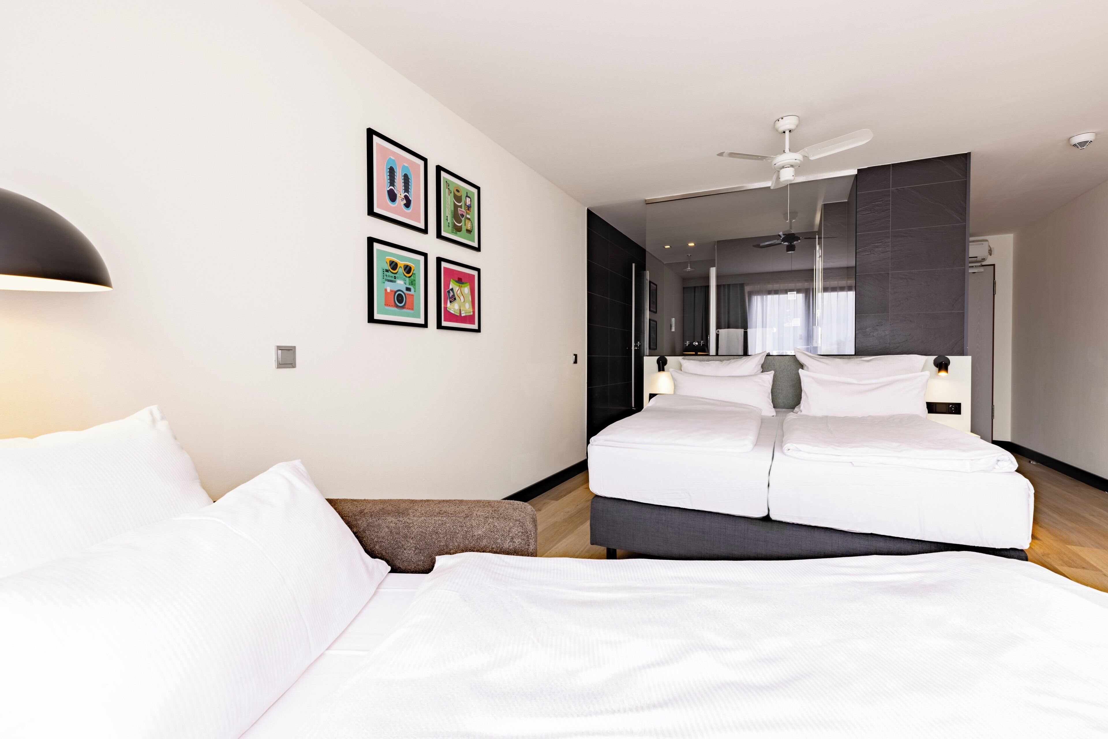 Une chambre d’hôtel moderne avec un grand lit, un petit coin salon, un ventilateur de plafond et des tableaux encadrés au mur.