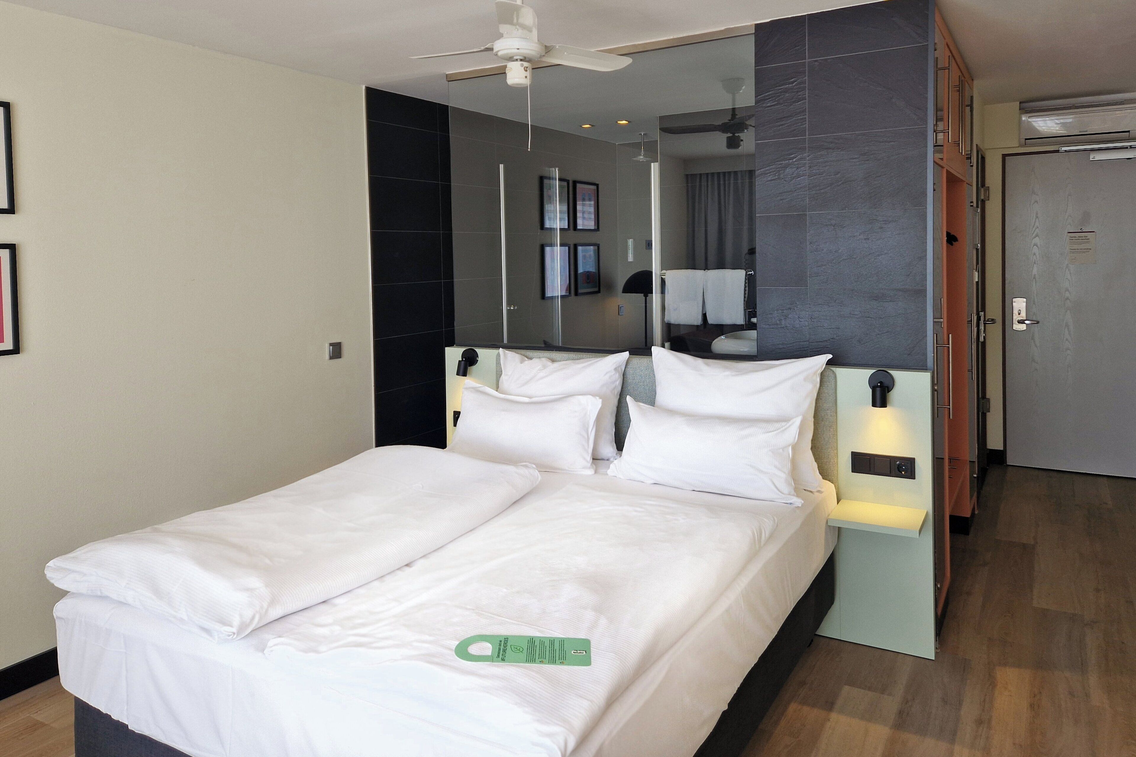 Ein Hotelzimmer mit einem Bett, einem Nachttisch mit Lampe, einem Deckenventilator, einem Spiegel und einer Tür.