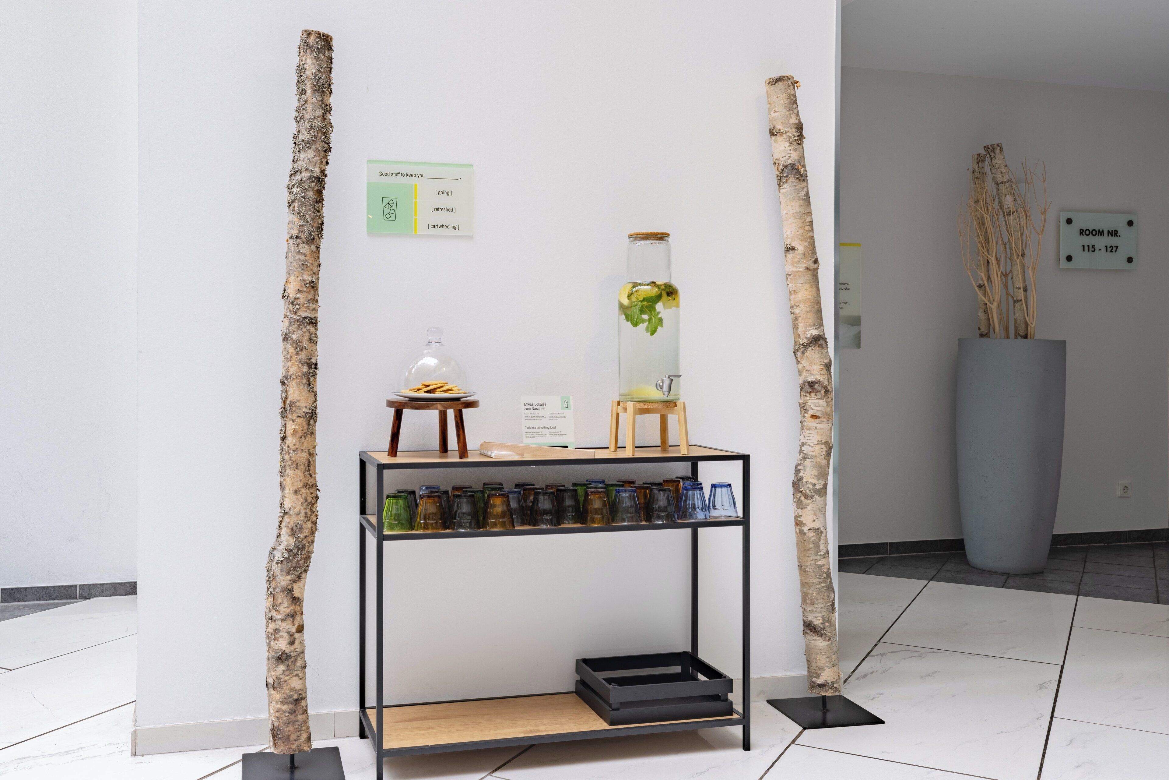 A modern interior with a minimalist design, featuring a wooden stand with glass bottles, a small table with a lamp, and a tall vase with greenery.