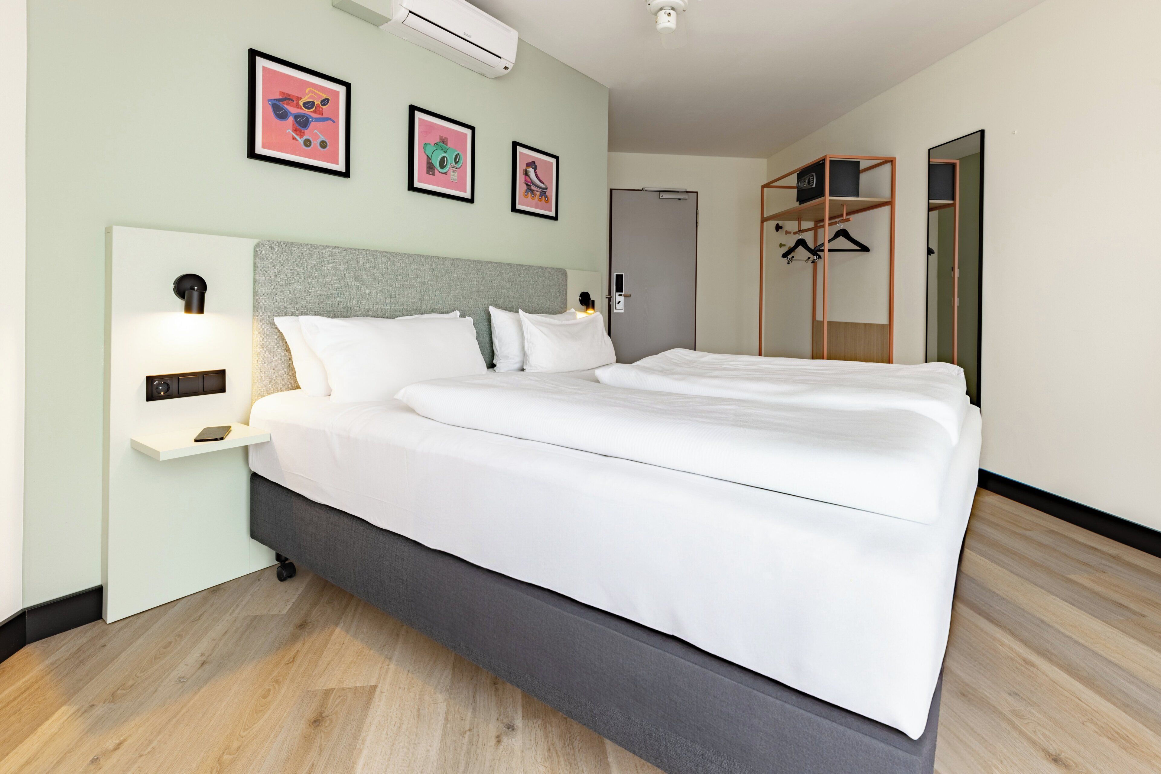 A modern hotel room with a large bed, a bedside table with a lamp, a wardrobe, and a mirror.