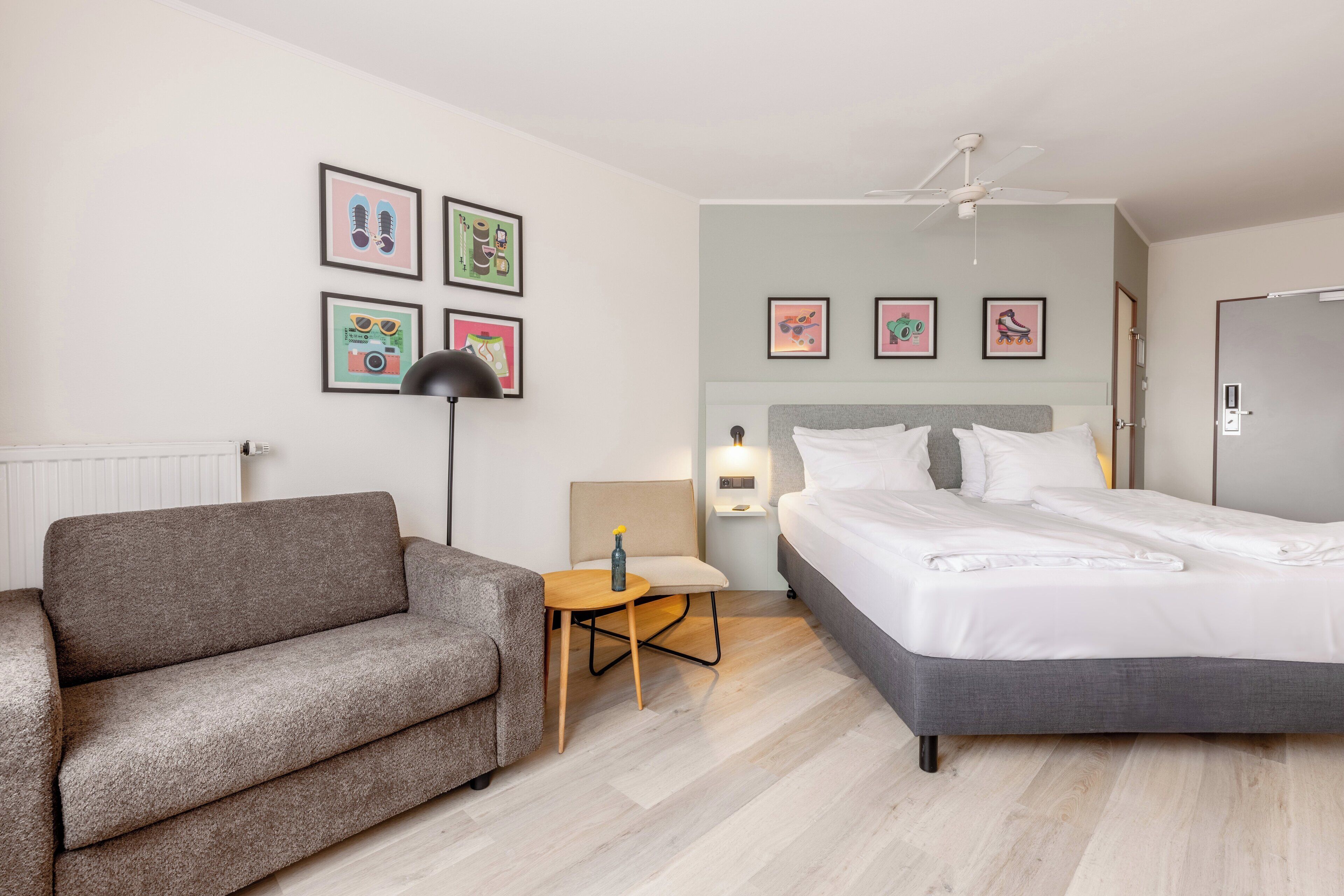 Una habitación de hotel moderna con un sofá gris, una mesita de madera, una cama con ropa blanca y un ventilador de techo.