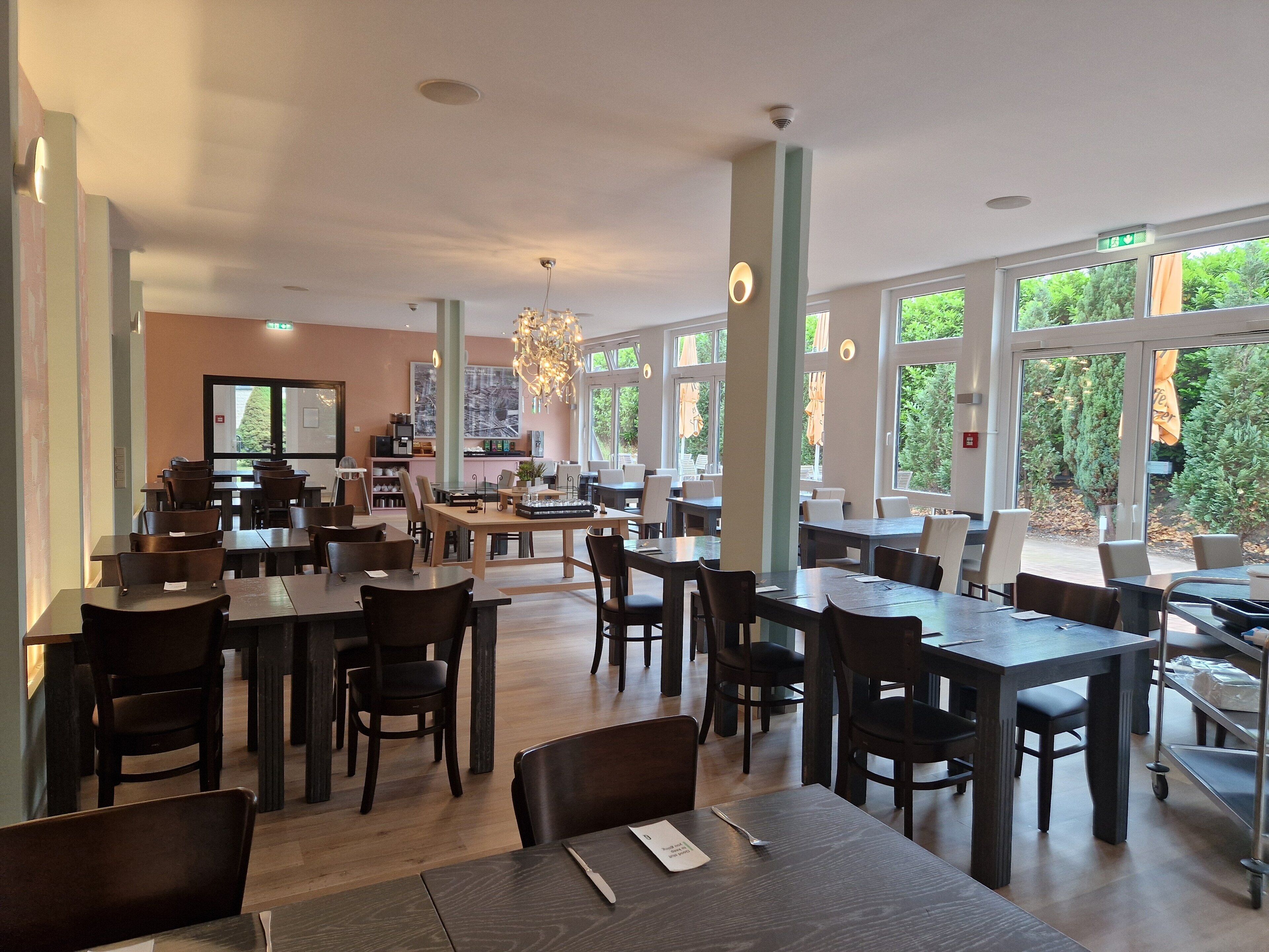A spacious dining area with multiple tables and chairs, large windows offering a view of greenery outside, and a central island with a chandelier.