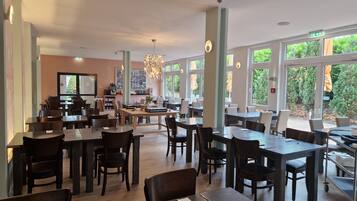 A spacious dining area with multiple tables and chairs, large windows offering a view of greenery outside, and a central island with a chandelier.