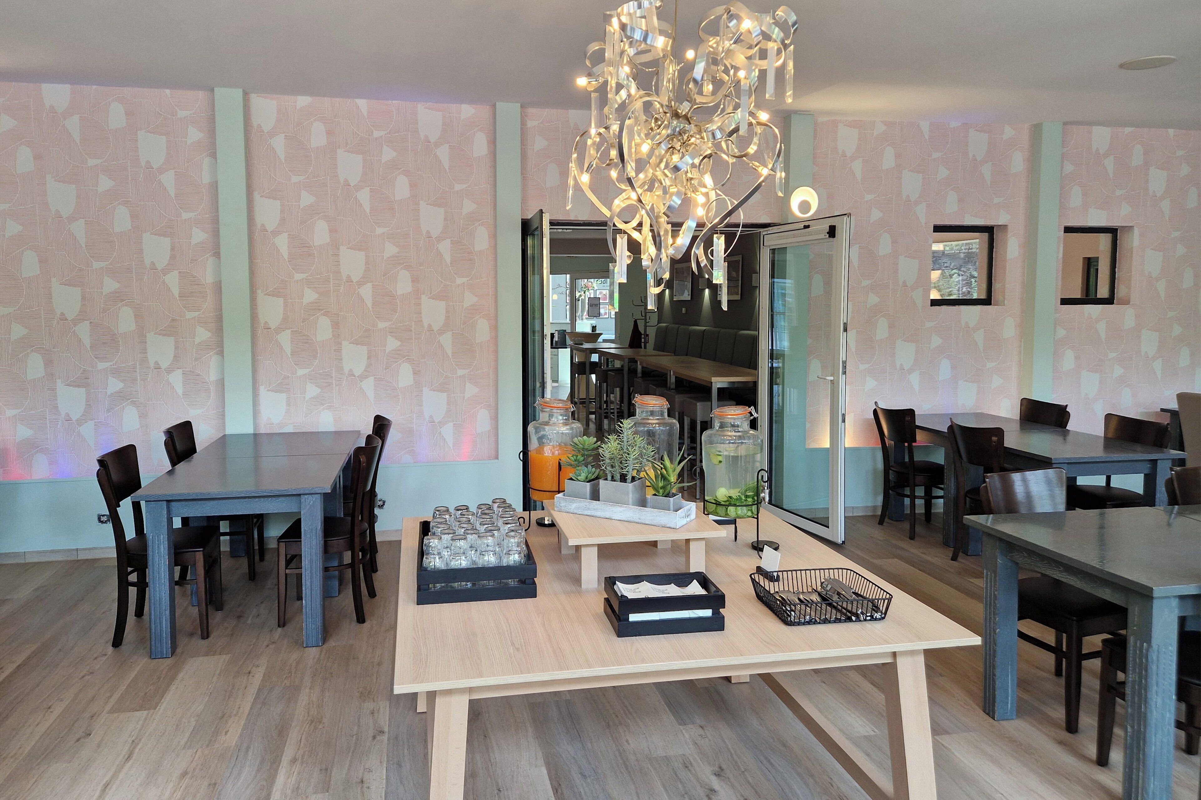 A modern dining area with a large table, chairs, a chandelier, and a kitchen in the background.