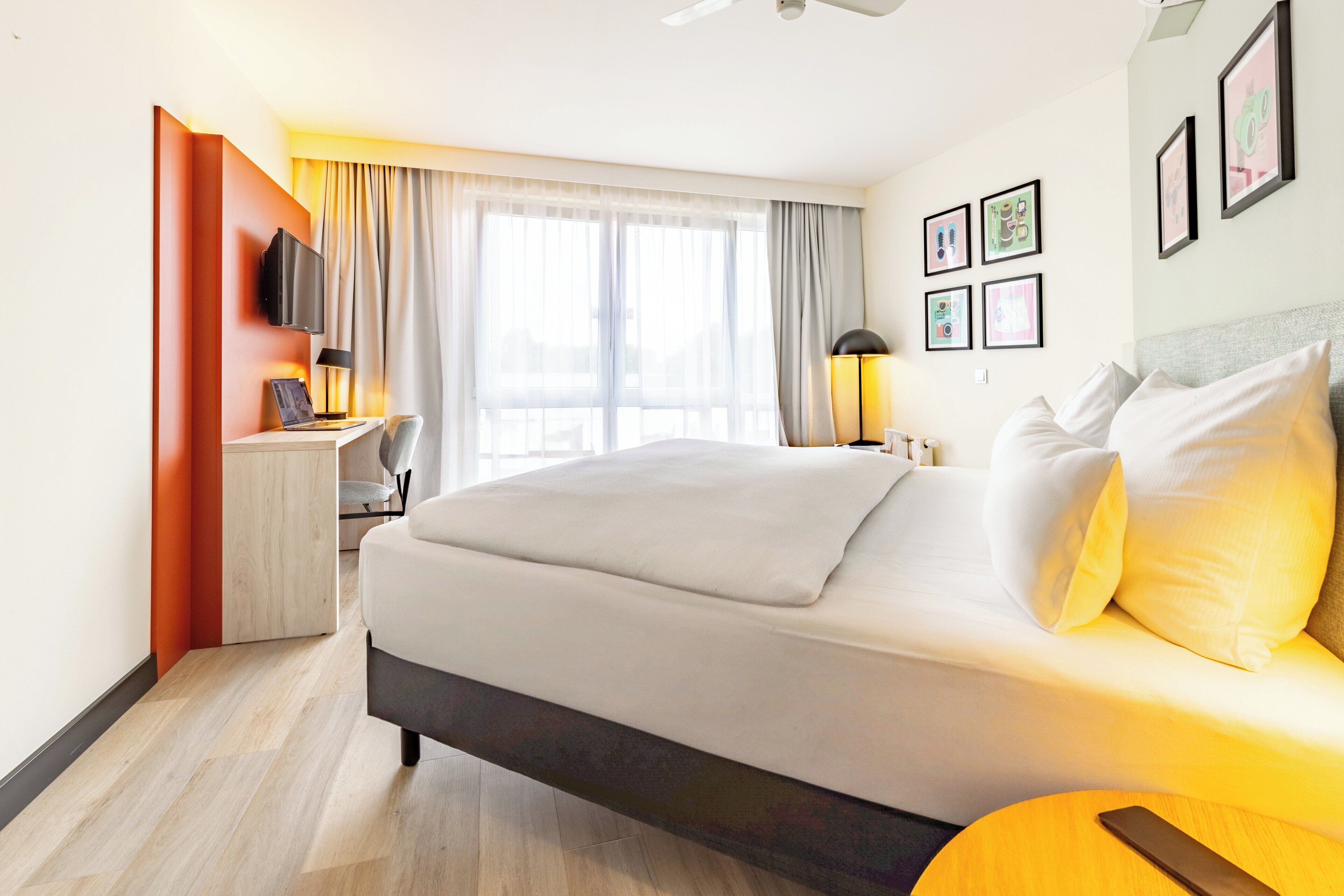 A hotel room with a large bed, a desk with a chair, a TV mounted on the wall, and a window with curtains.