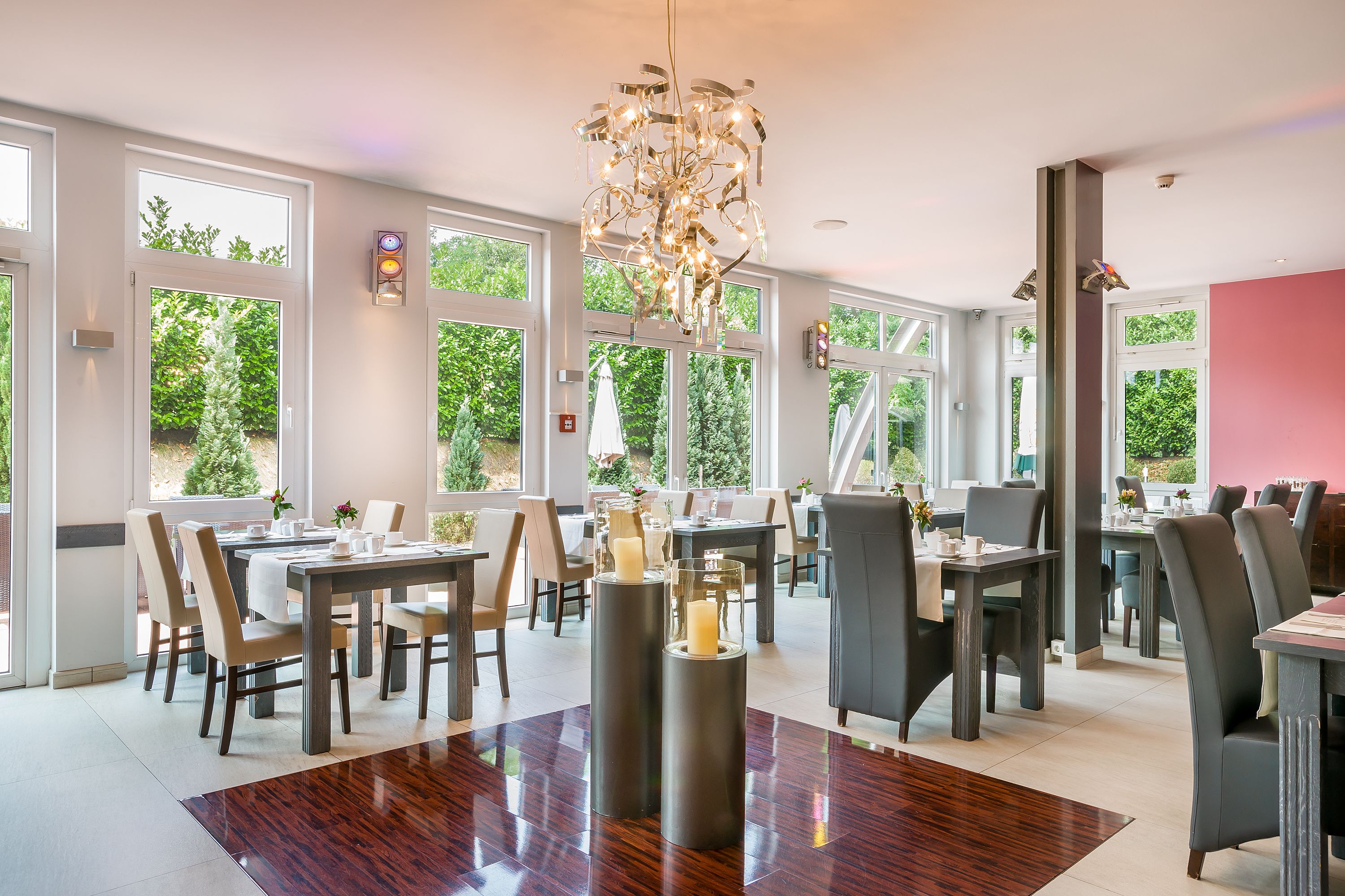 A spacious dining area with multiple tables and chairs, large windows offering views of greenery, and a modern chandelier.