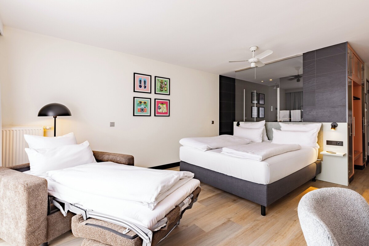 A modern hotel room with a bed, a sofa, a chair, a ceiling fan, and framed pictures on the wall.