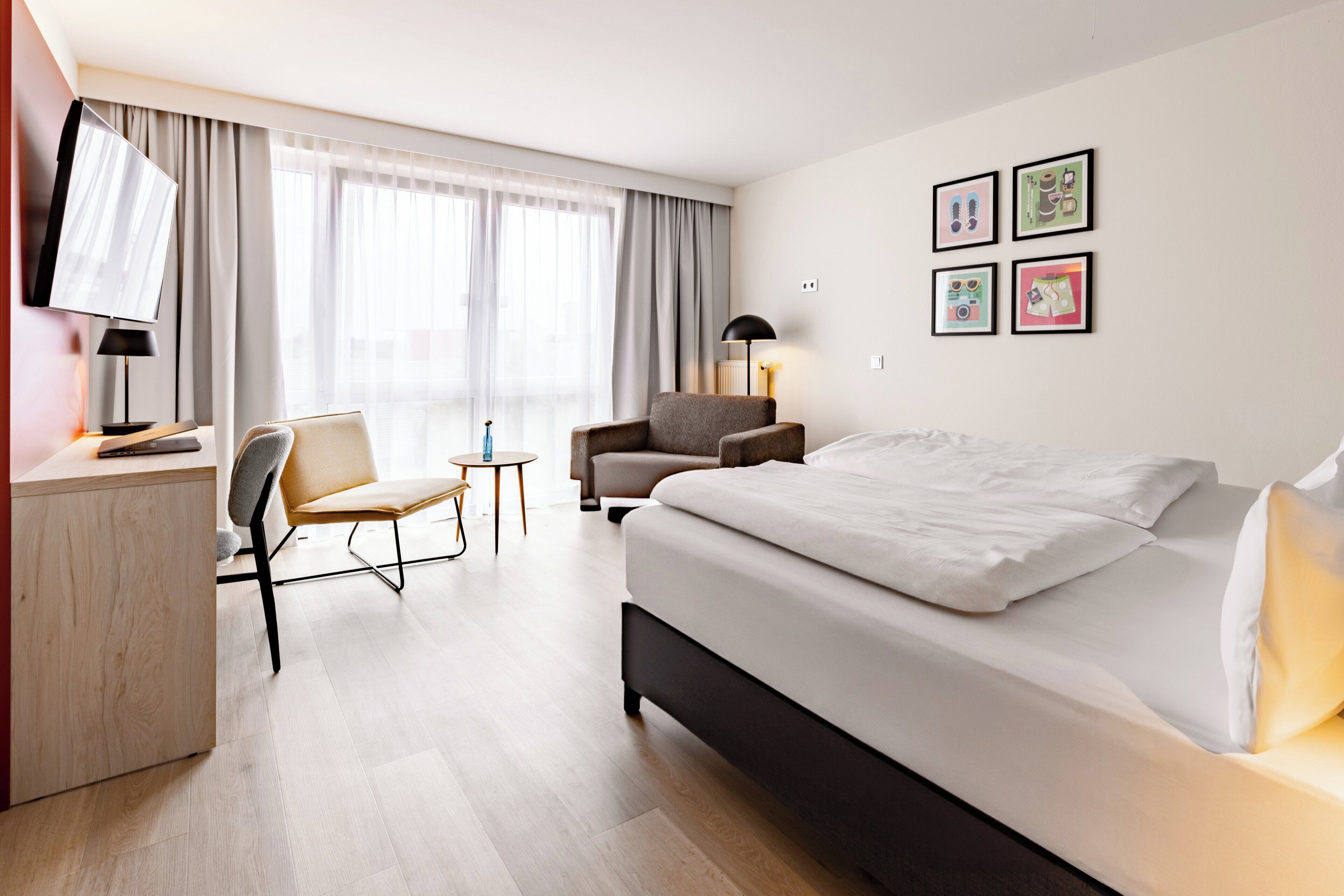 A modern hotel room with a large bed, a desk, a chair, a small table, and a TV. There are curtains, a lamp, and framed pictures on the wall.
