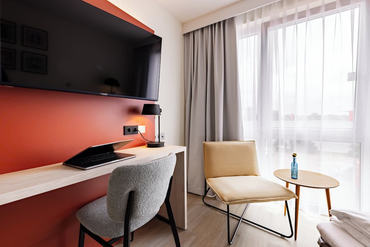 A modern hotel room with a desk, laptop, chair, and a small table with a bottle.