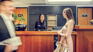 A hotel reception area with a receptionist, a man holding papers, and a woman in a dress. - Mercure Hotel München Altstadt (Munich)