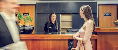 A hotel reception area with a receptionist, a man holding papers, and a woman in a dress.