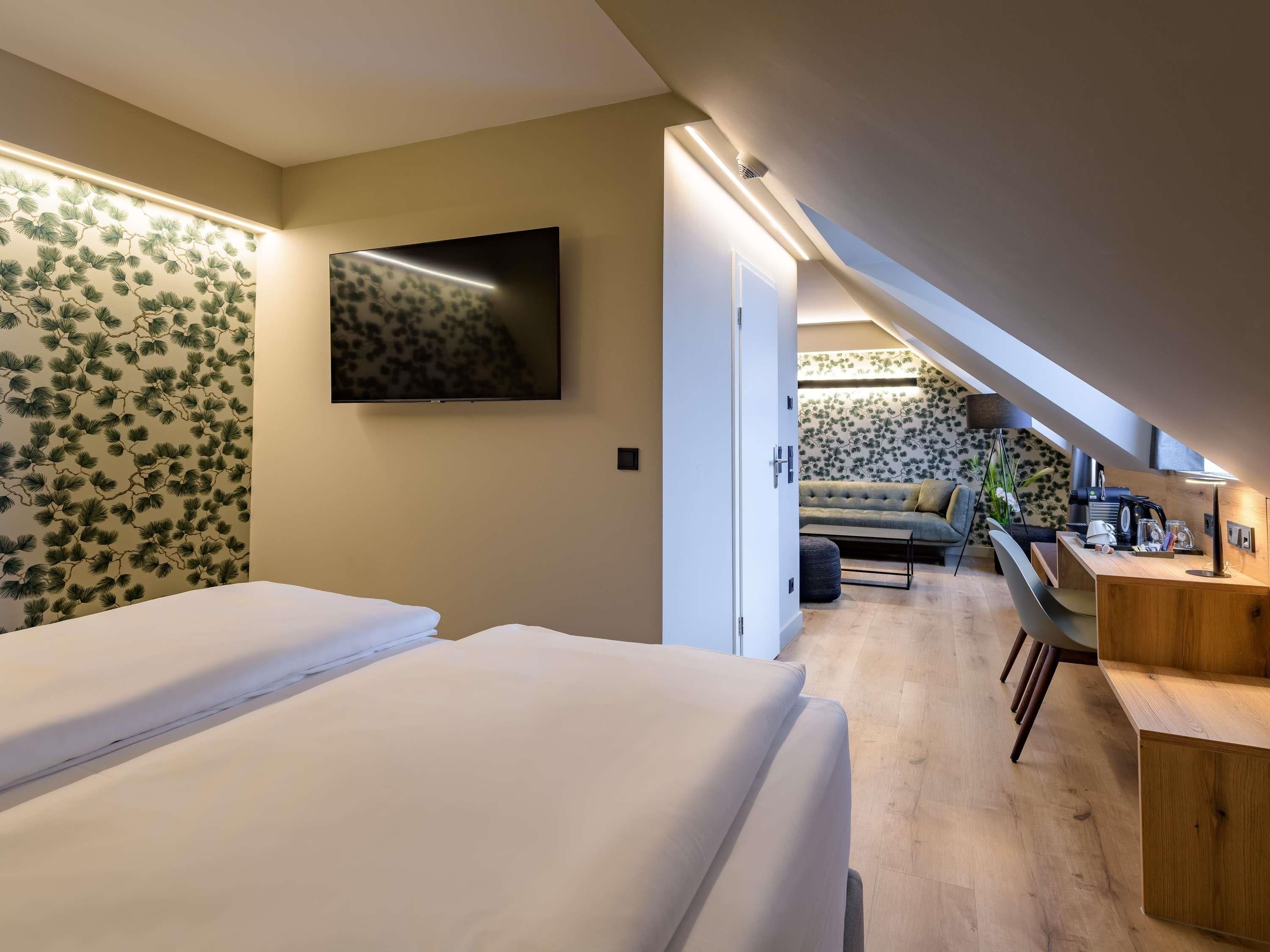 Un dormitorio con dos camas, un televisor, un escritorio con silla, y una escalera que lleva a una sala de estar con sofá y cafetera.