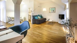 A modern living room with a sofa, a dining area with chairs, a TV, and a lamp. - Mercure Hotel München Altstadt (Munich)
