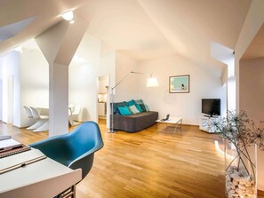 A modern living room with a sofa, a dining area with chairs, a TV, and a lamp. - Mercure Hotel München Altstadt (Munich)