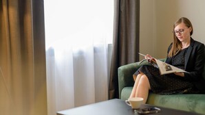 A person sitting on a green sofa in a room with a window, reading a magazine. - Mercure Hotel München Altstadt (Munich)