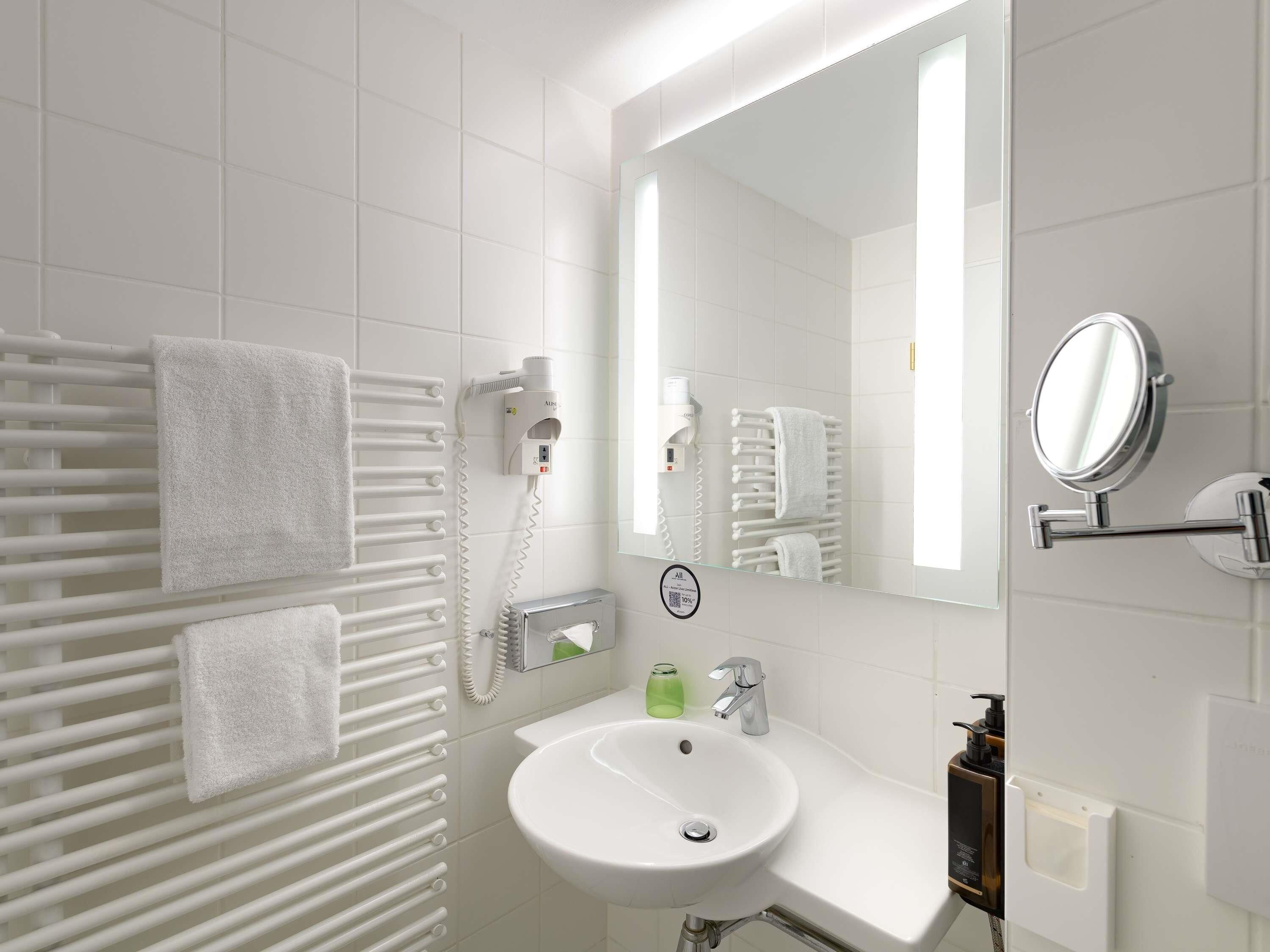 A modern bathroom with a white sink, a mirror, a towel rack, a hairdryer, and a soap dispenser.