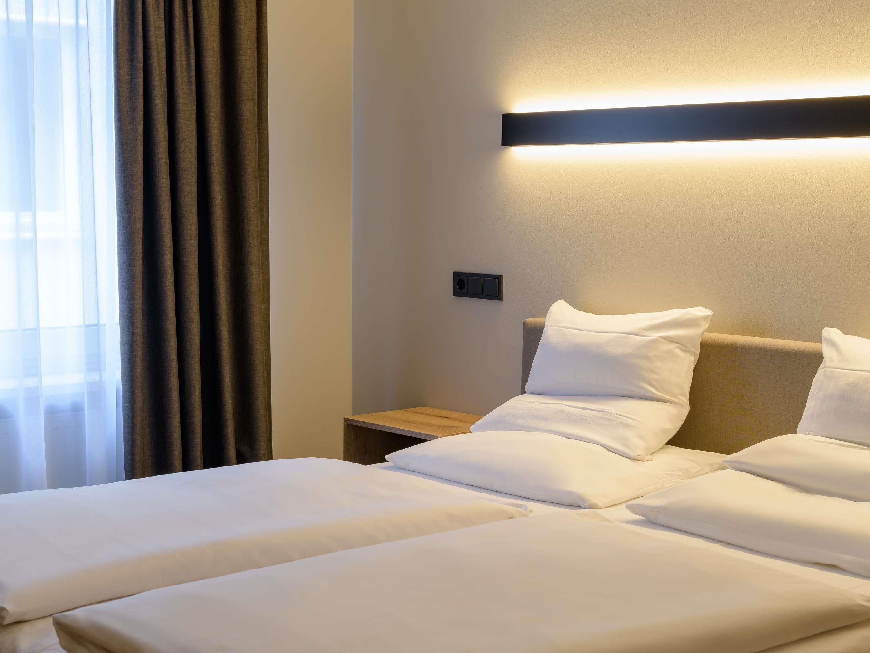 Une chambre d’hôtel avec deux lits, une table de chevet, une lampe fixée au mur et une fenêtre avec des rideaux.