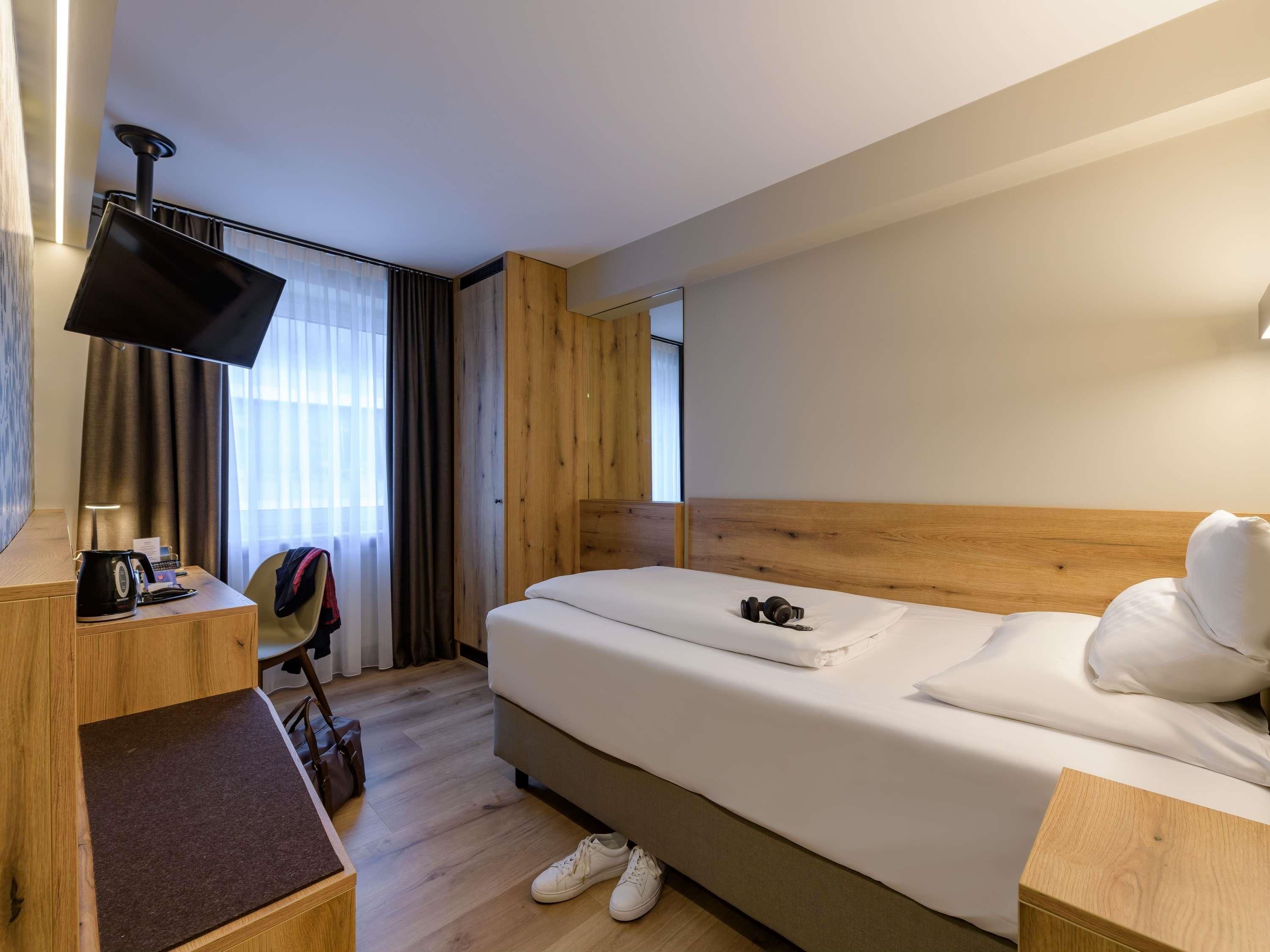 A hotel room with a bed, a desk with a flat-screen TV, a chair, and a suitcase on the floor.