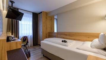 A hotel room with a bed, a desk with a flat-screen TV, a chair, and a suitcase on the floor.